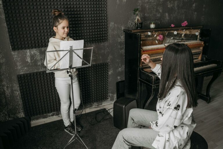 A young girl receiving vocal training from a woman near a piano in a music studio.