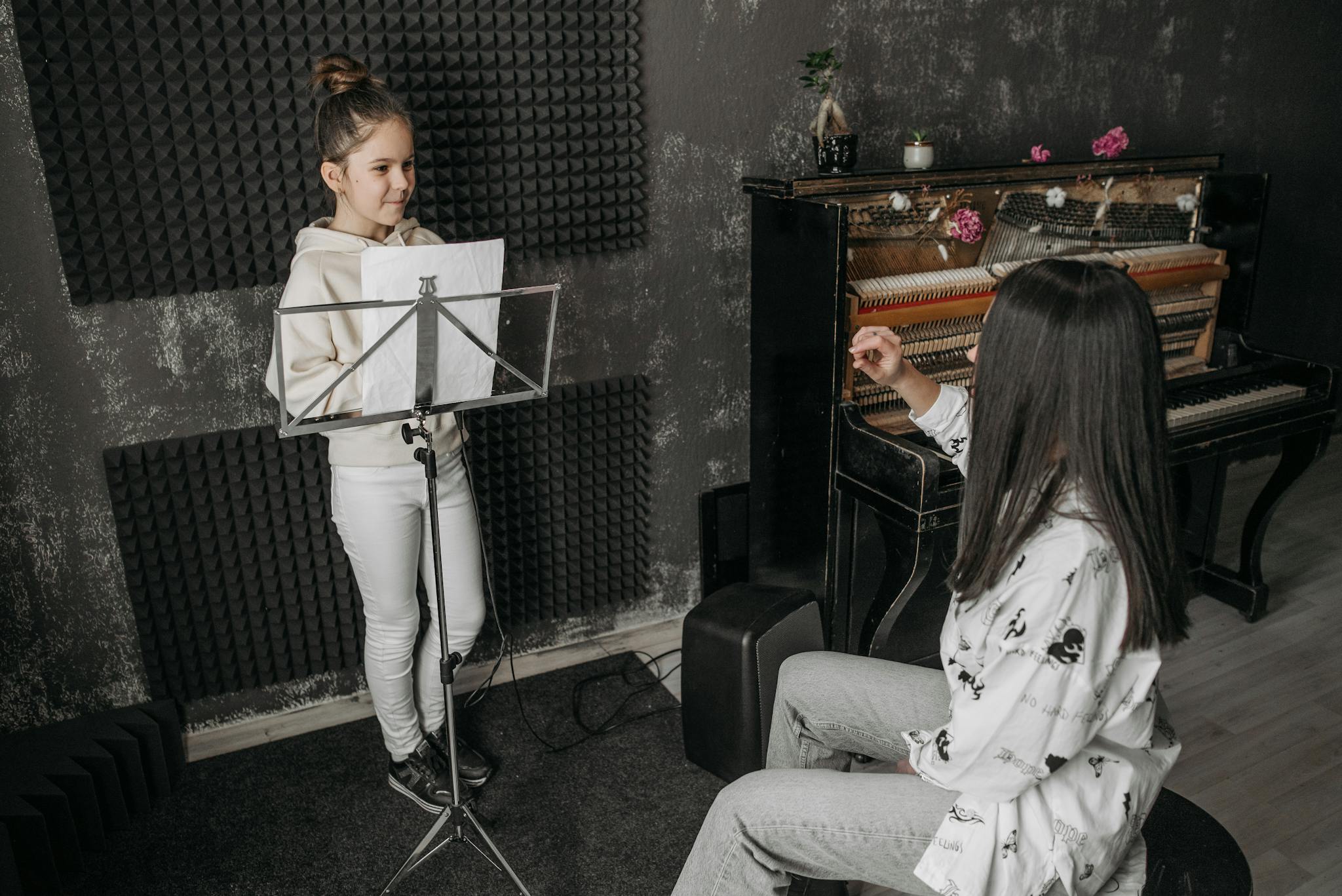 A young girl receiving vocal training from a woman near a piano in a music studio.