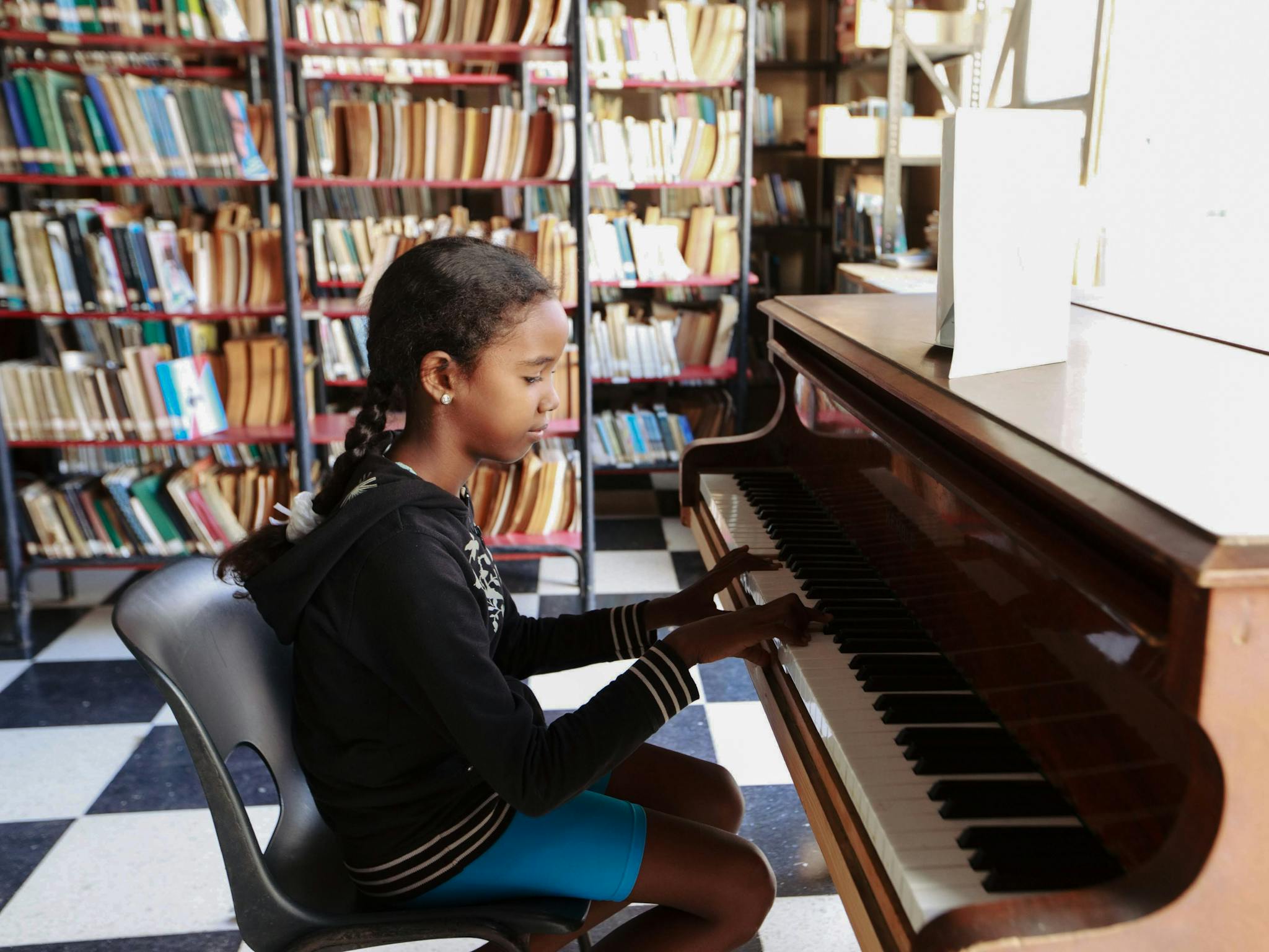 A young girl sits at a piano in a library, playing music in a bright and inviting space.