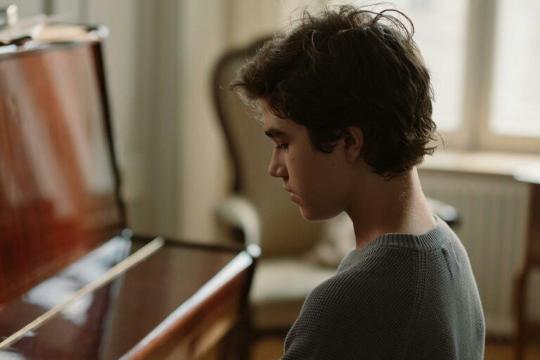 Young boy sitting by a piano, side view, captured indoors in soft lighting.