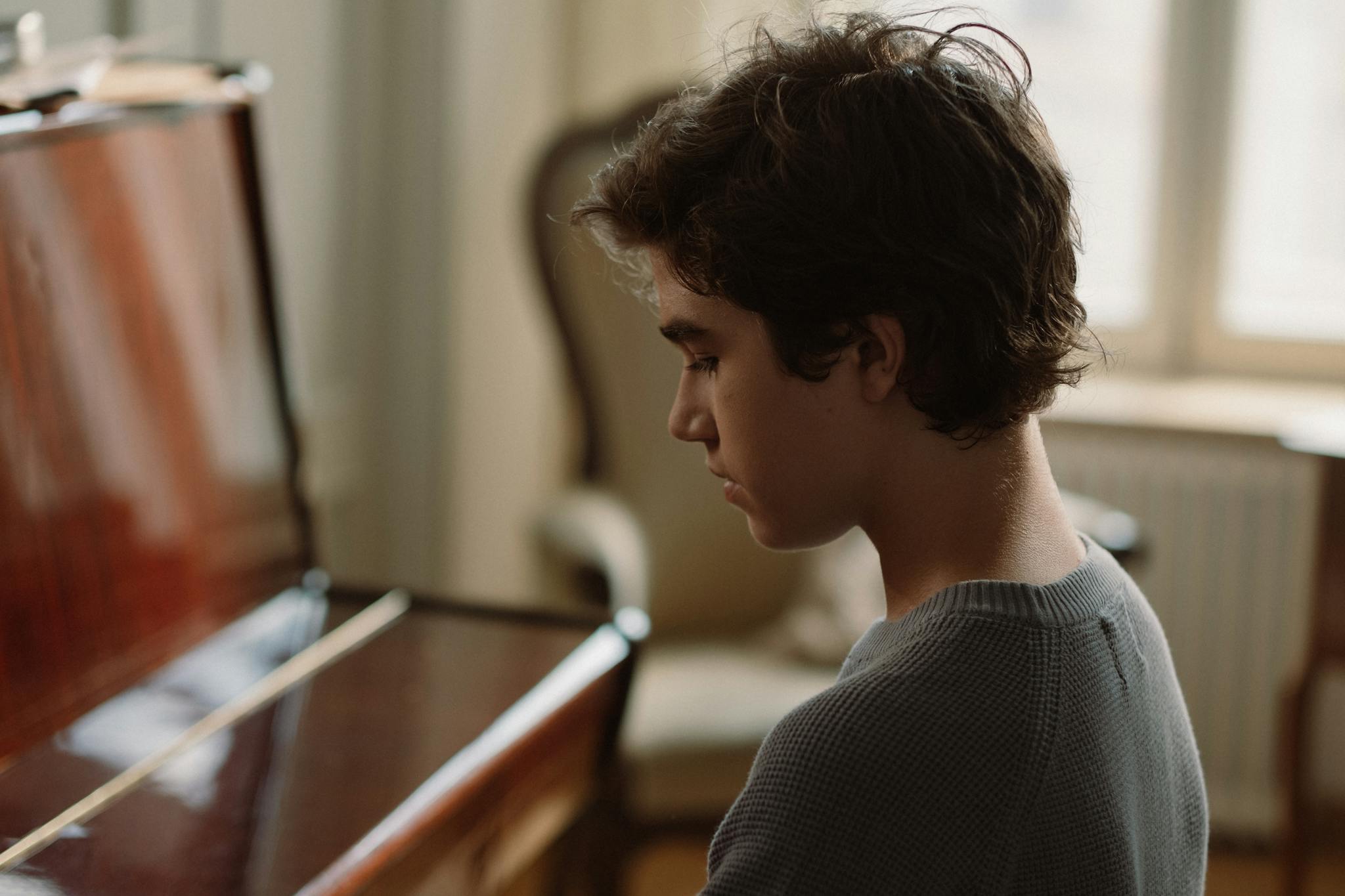 Young boy sitting by a piano, side view, captured indoors in soft lighting.